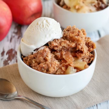 Square image of apple crumble in a white bowl topped with vanilla ice cream with more servings in the background.