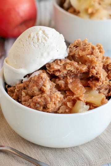 Square image of apple crumble in a white bowl topped with vanilla ice cream with more servings in the background.