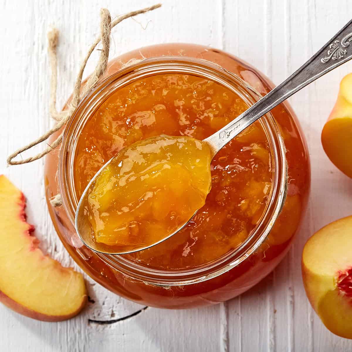 A jar of Peach Jam with a metal spoon resting on top, surrounded by fresh peach slices on a white wooden surface.