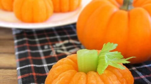 Small pumpkin-shaped snacks made from peeled mandarin oranges with celery stalks as stems, these Healthy Halloween Treats are arranged on a plaid cloth and a plate.
