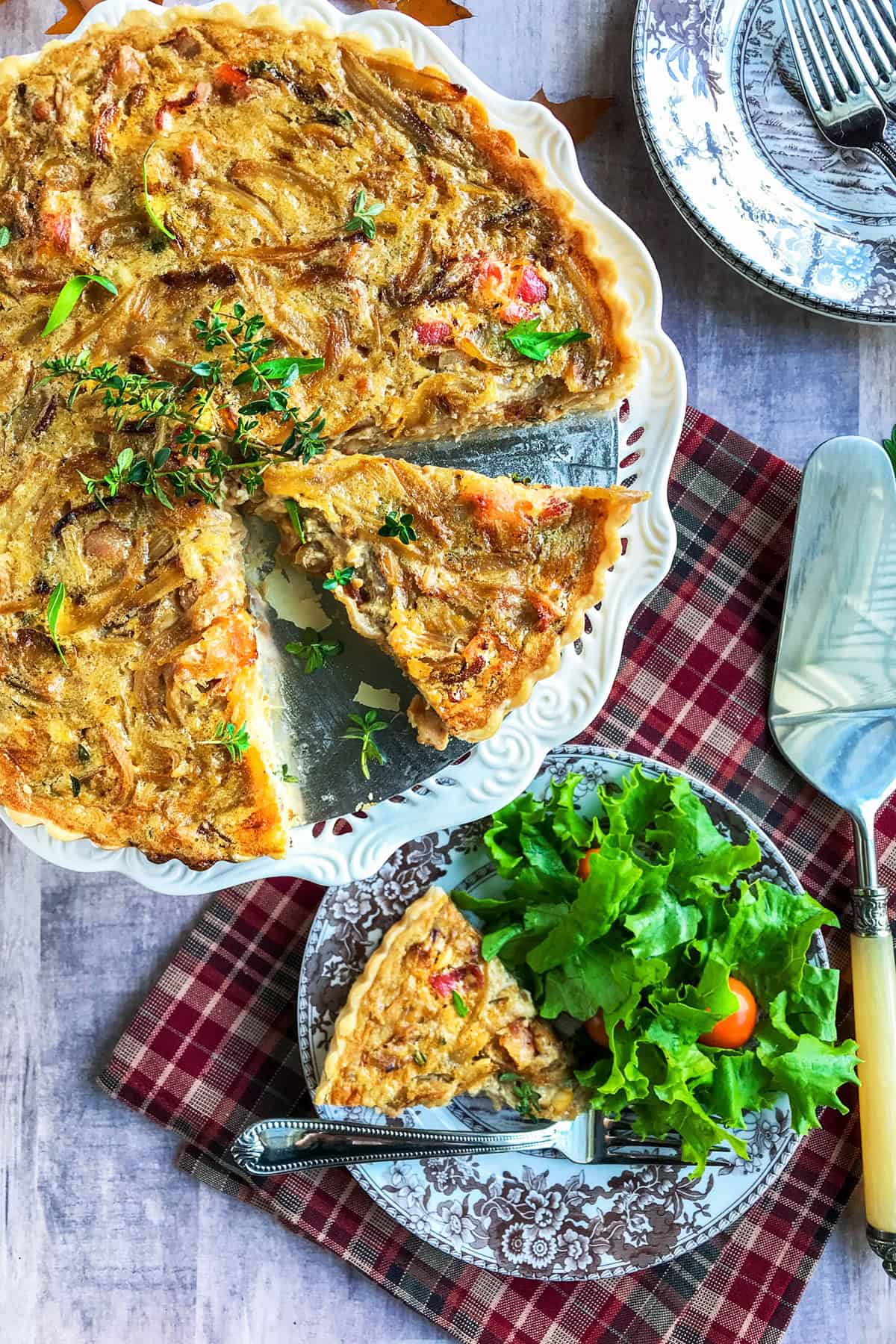 Sliced onion tart with fresh herbs as garish, a plaid linen underneath and a nearby plate with a slice, fork, and side salad.