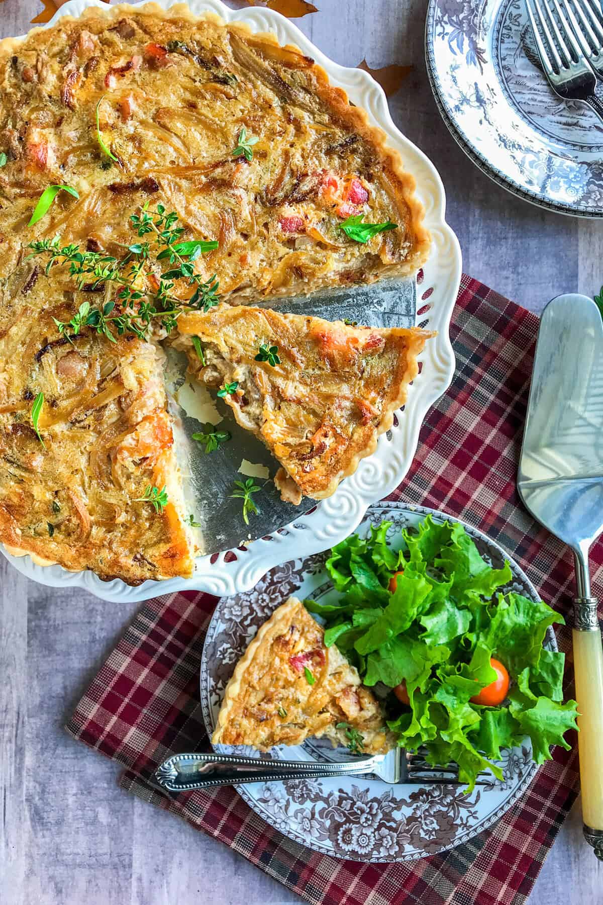 A baked quiche with a golden-brown top is served on a white platter, with one creamed onion slice removed and placed on a plate alongside a fresh green salad. A pie server and utensils are nearby on a red checkered cloth.