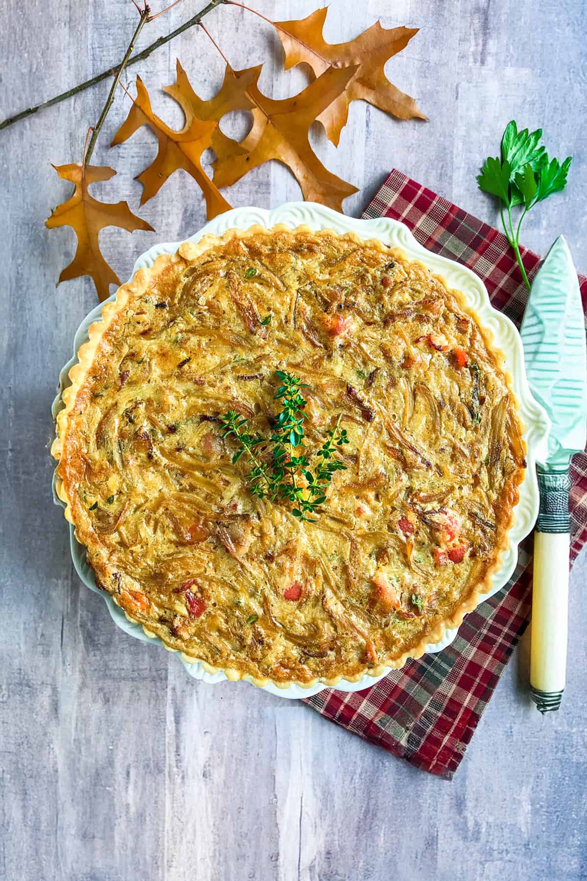Unsliced French Onion Tart with fresh thyme garnish on a white serving stand with fall leaves in the background.