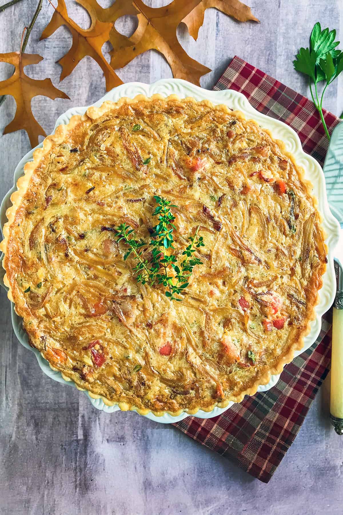 A golden-brown quiche with creamed onion, garnished with fresh thyme, sits on a white plate atop a red plaid napkin. Autumn leaves, parsley, and a knife are arranged around it on a gray tabletop.