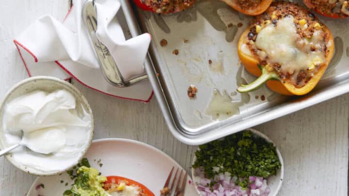 Flavor packed comfort food dinners: Baked stuffed bell peppers with melted cheese in a tray, served with sour cream, guacamole, chopped red onion, and herbs on the side.