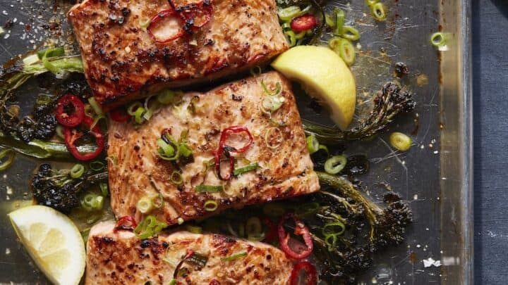 Four pieces of cooked salmon topped with sliced green onions and red chilies on a baking sheet with roasted greens and lemon wedges—perfect for cozy October Savory Dinners.