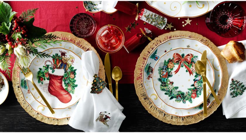 Festive table setting with Christmas-themed plates, gold flatware, holiday napkins, a red tablecloth, drinks perfect for a Christmas Tea, and greenery decor.