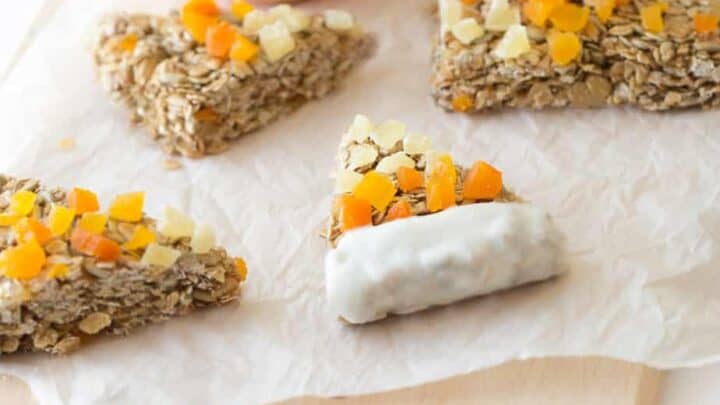 Four triangular oat bars topped with chopped dried fruit on parchment paper, one partially dipped in white coating, make festive Healthy Halloween Treats, served with a small orange bowl of white yogurt in the background.