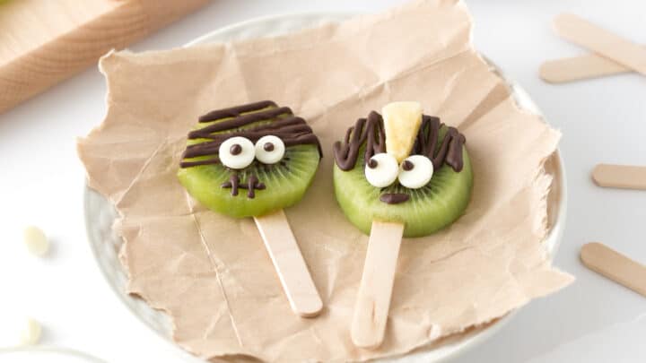 Two kiwi slices on popsicle sticks decorated with candy eyes and chocolate drizzle, styled to look like faces, make fun and Healthy Halloween Treats on a crumpled piece of parchment paper.