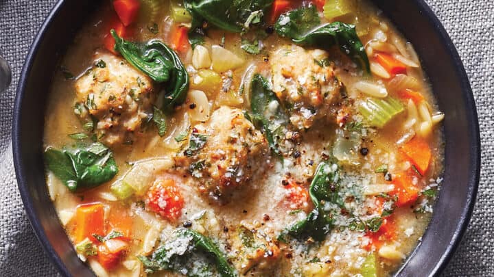 A cozy November Dinners favorite: a bowl of soup with meatballs, orzo pasta, spinach, carrots, celery, and broth, garnished with grated cheese and fresh herbs.