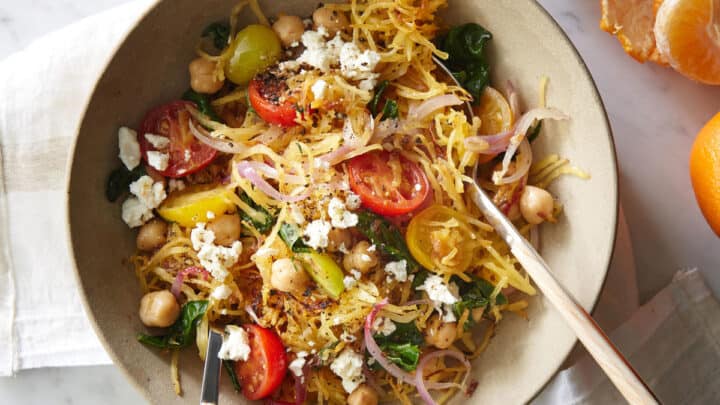 A bowl of spaghetti squash salad with cherry tomatoes, chickpeas, spinach, red onion, and crumbled feta cheese—perfect for cozy November Dinners—served with utensils on a white napkin.