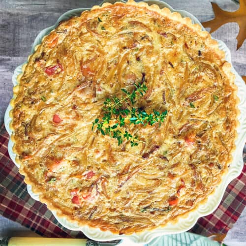 A baked quiche with a golden, textured top sits on a white plate, garnished with fresh herbs and hints of creamed onion. The plate rests on a red plaid cloth, with autumn leaves and a rolling pin nearby.