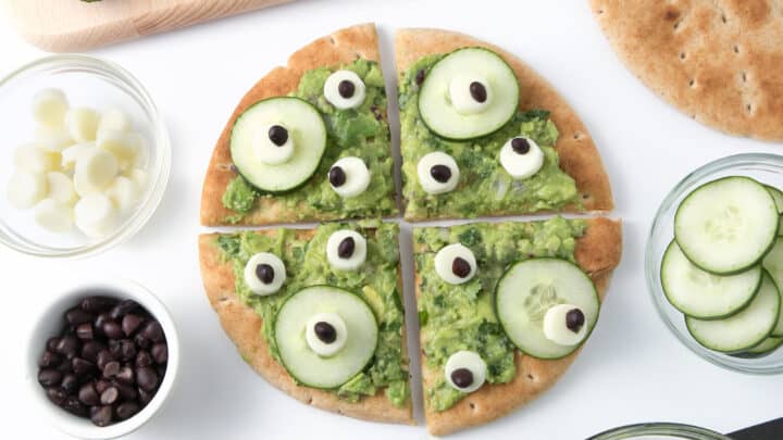Round flatbread with guacamole, cucumber slices, and olive-topped cheese circles arranged to look like eyes—an adorable addition to your Healthy Halloween Treats lineup, surrounded by fresh ingredients on a white surface.