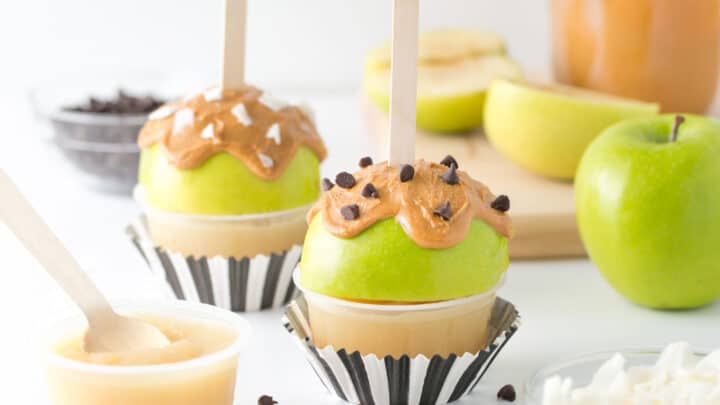 Two green apples with popsicle sticks, topped with peanut butter and mini chocolate chips, sit in striped cups; applesauce, apple slices, and toppings are arranged around them for festive Healthy Halloween Treats.