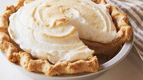 A hand pipes white whipped cream onto the edge of a Thanksgiving Pie-inspired red fruit tart on a marble surface.