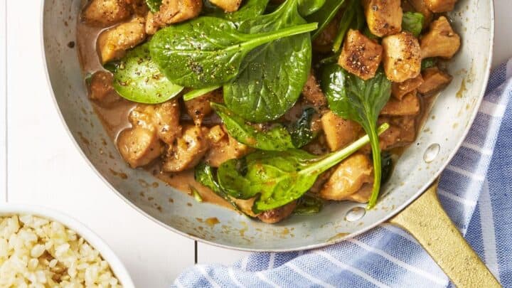 A pan with cooked chicken pieces and spinach in sauce, perfect for healthy fall dinners, sits next to a bowl of brown rice on a blue and white striped cloth atop a white surface.
