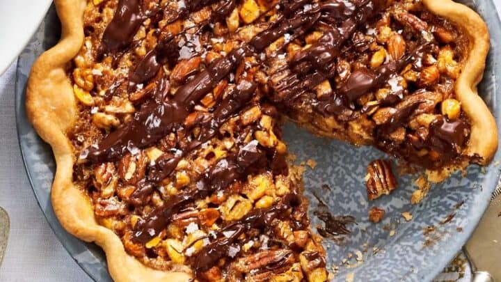 A chocolate-drizzled Thanksgiving Pie sits in a metal pan with one slice removed, revealing the nut filling and fluted crust; a pie server rests nearby.