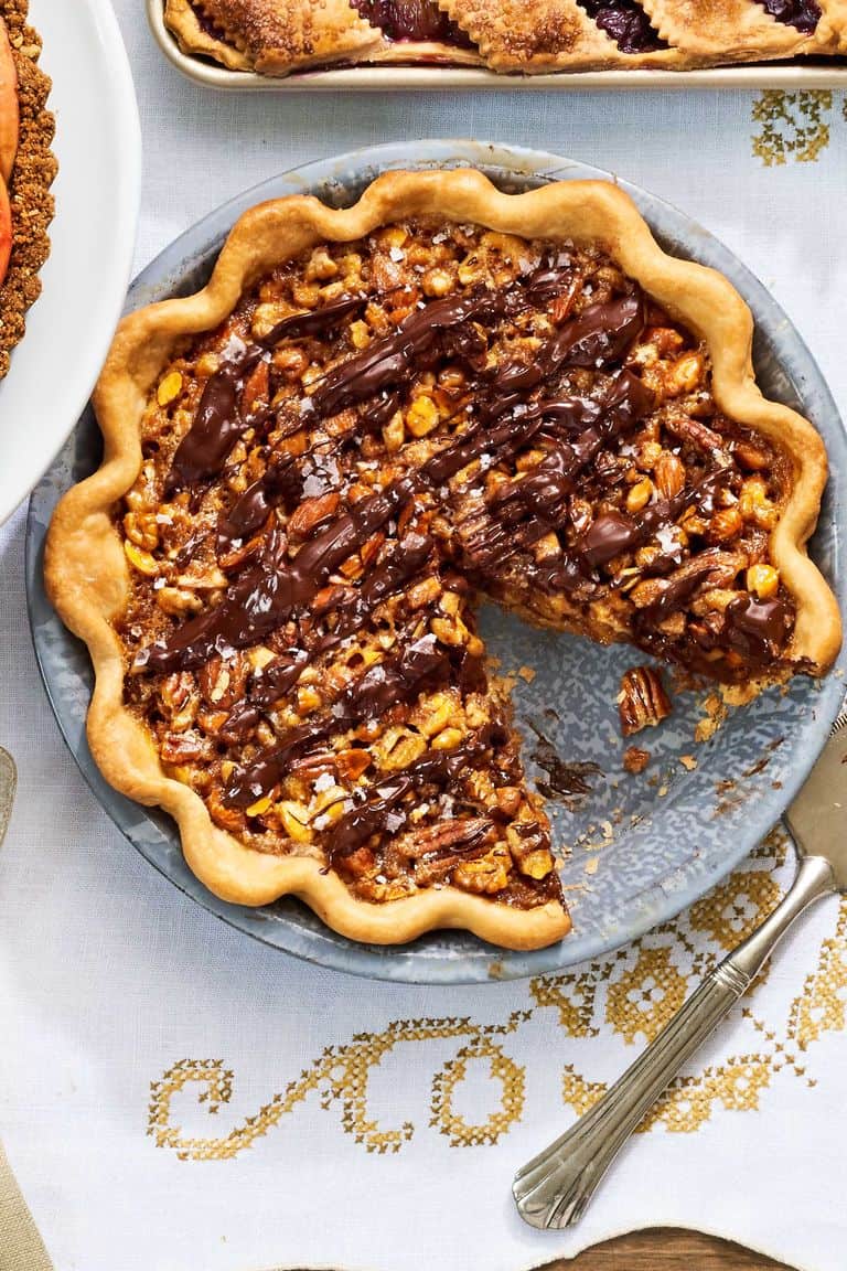 A chocolate-drizzled Thanksgiving Pie sits in a metal pan with one slice removed, revealing the nut filling and fluted crust; a pie server rests nearby.