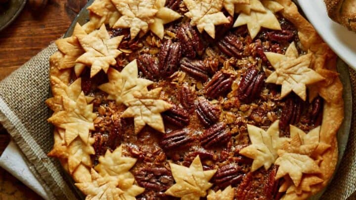 Thanksgiving Pie: Pecan pie topped with decorative leaf-shaped pastry, displayed on a rustic wooden table with plates and a bread loaf nearby.