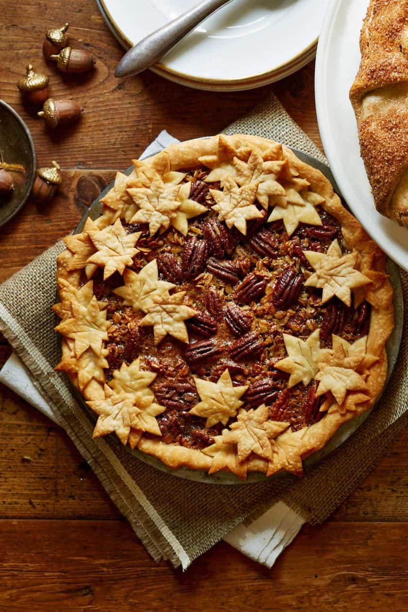Thanksgiving Pie: Pecan pie topped with decorative leaf-shaped pastry, displayed on a rustic wooden table with plates and a bread loaf nearby.