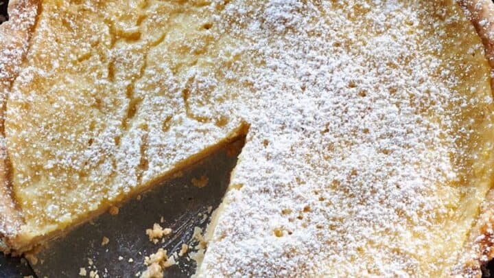 A round Thanksgiving Pie with a flaky crust and powdered sugar on top, missing one slice, sits in a metal tart pan on a wooden surface.