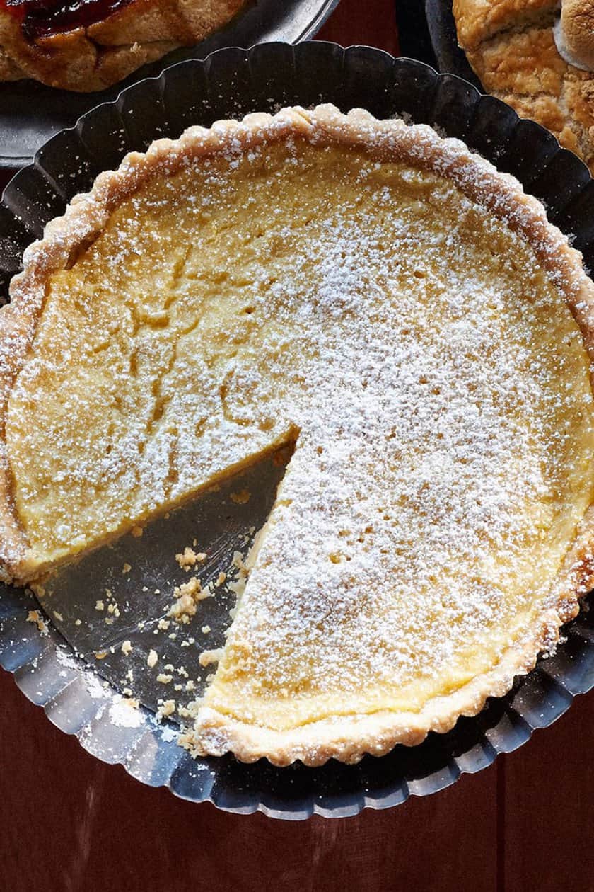 A round Thanksgiving Pie with a flaky crust and powdered sugar on top, missing one slice, sits in a metal tart pan on a wooden surface.