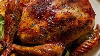 Roast whole turkey garnished with herbs and lemon slices on a white platter, served with a carving knife—a classic centerpiece for Turkey recipes for Thanksgiving.