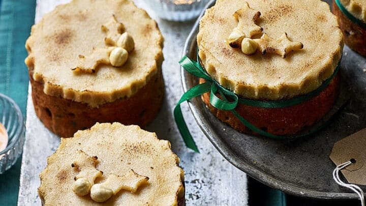 Three round festive cakes with decorative pastry toppings, one tied with green ribbon, displayed on a tray—perfect for celebrating Stir-Up Sunday—with labels, twine, candles, and string lights in the background.