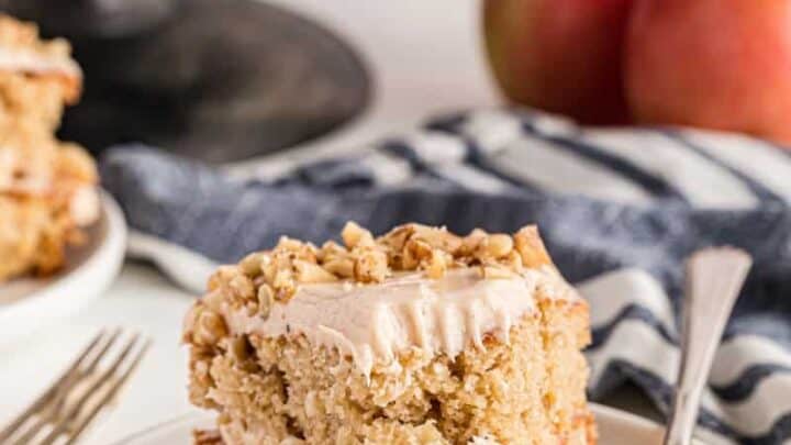 A slice of apple cake with frosting and walnuts sits on a white plate with a fork—an inviting addition to your Thanksgiving Desserts table. Apples and another cake are in the background.