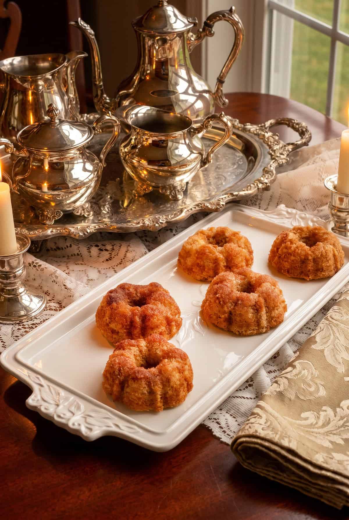 A white platter with five cinnamon teacakes sits on a lace tablecloth, next to ornate silver tea sets, candles, and a window-a scene reminiscent of Dolley Madison's elegant gatherings.