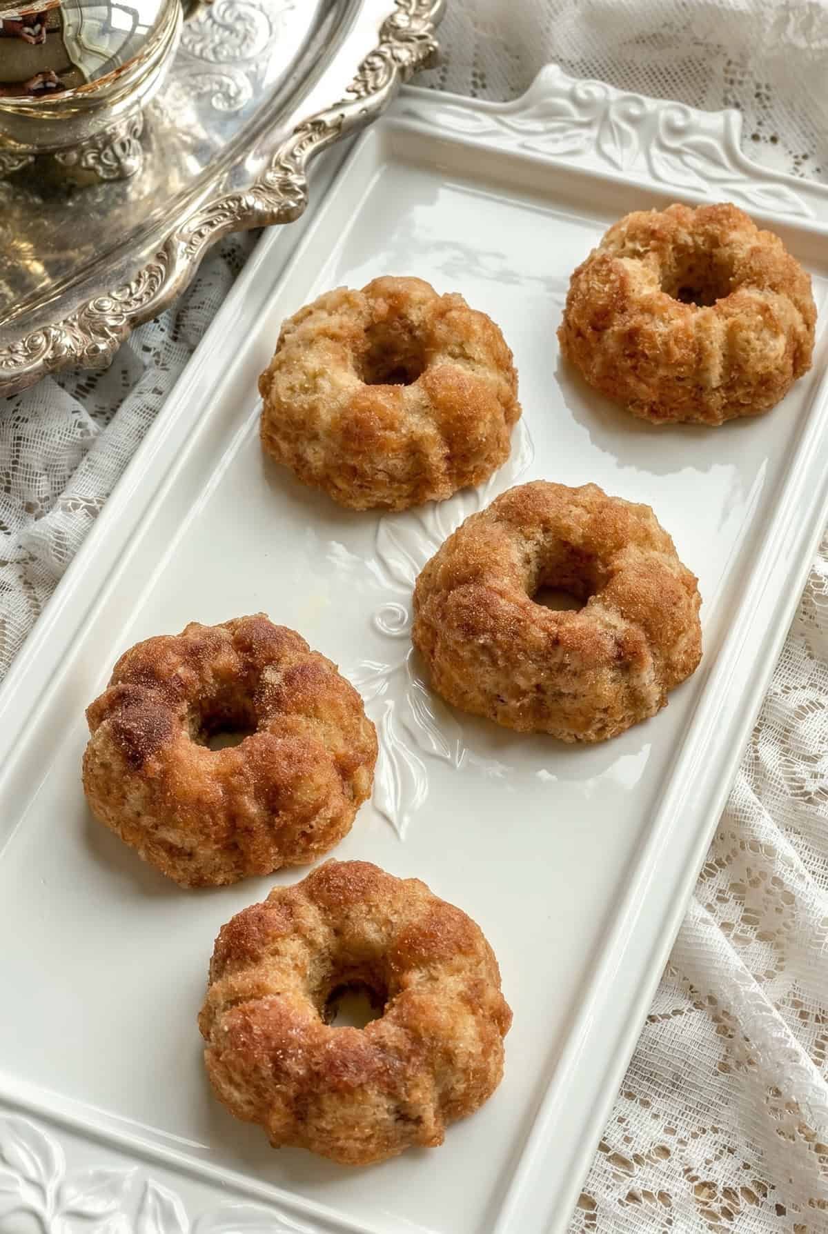 Five cinnamon teacakes, reminiscent of Dolley Madison's elegant gatherings, are arranged on a rectangular white tray placed on a lace tablecloth beside a silver tray.