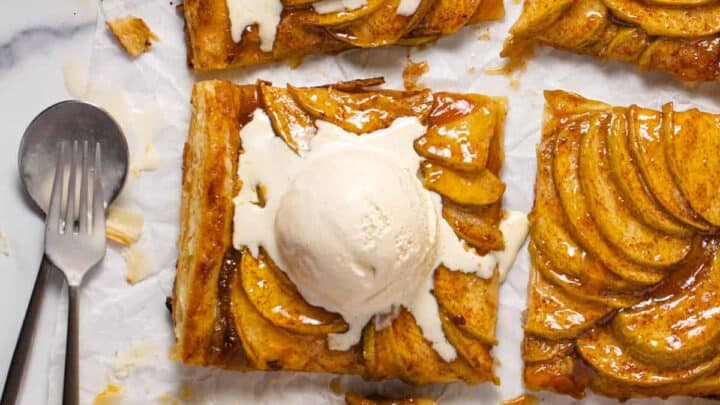 Six rectangular slices of apple tart topped with vanilla ice cream are arranged on parchment paper, making a tempting addition to your Thanksgiving desserts. A cup of sauce, fork, and spoon sit nearby, ready for serving.