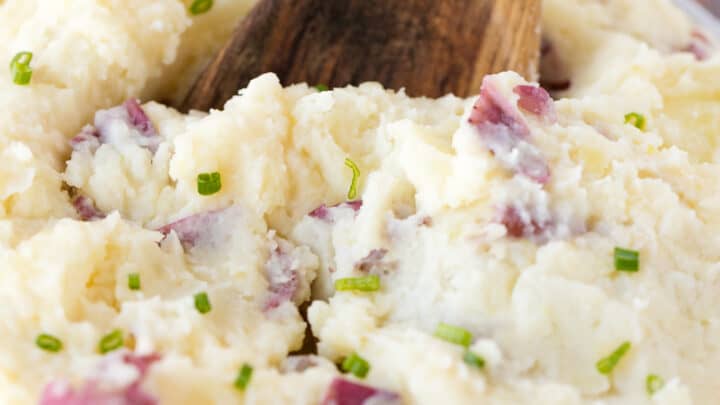 A wooden spoon in a bowl of creamy mashed potatoes with red potato skins and chopped chives, perfect for your favorite mashed potatoes recipes.