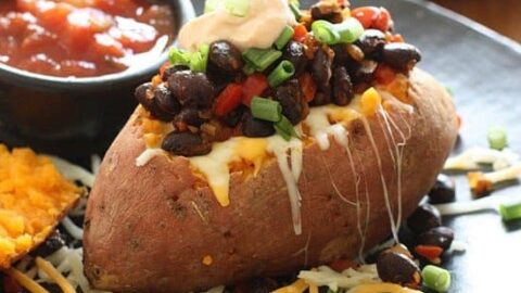 A healthy 20 minute dinner, this baked sweet potato is topped with black beans, shredded cheese, green onions, sour cream, and salsa, served on a black plate with extra salsa and cheese.