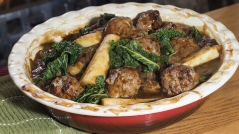 A ceramic dish filled with meatballs, greens, and root vegetables in a brown gravy, placed on a wooden surface—perfect comfort food for St Andrew's Day recipes.