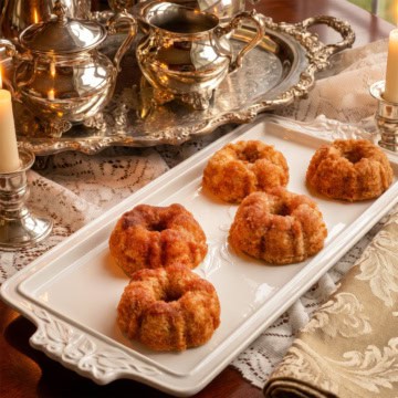 Five small cinnamon teacakes are arranged on a rectangular white platter next to a decorative silver tea set, candles, and a folded ornate napkin on a lace-covered table&mdash;perfect for a Dolley Madison-inspired gathering.