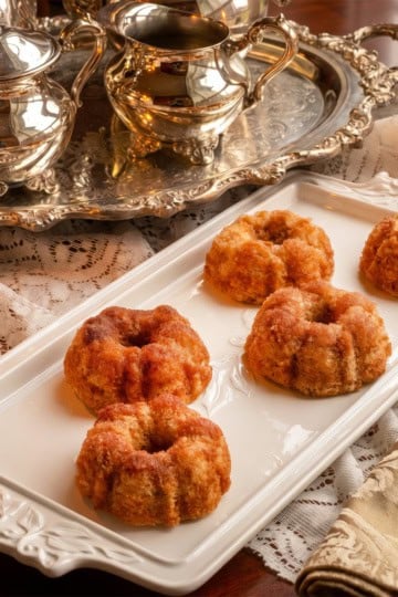 Five small cinnamon teacakes are arranged on a rectangular white platter next to a decorative silver tea set, candles, and a folded ornate napkin on a lace-covered table-perfect for a Dolley Madison-inspired gathering.