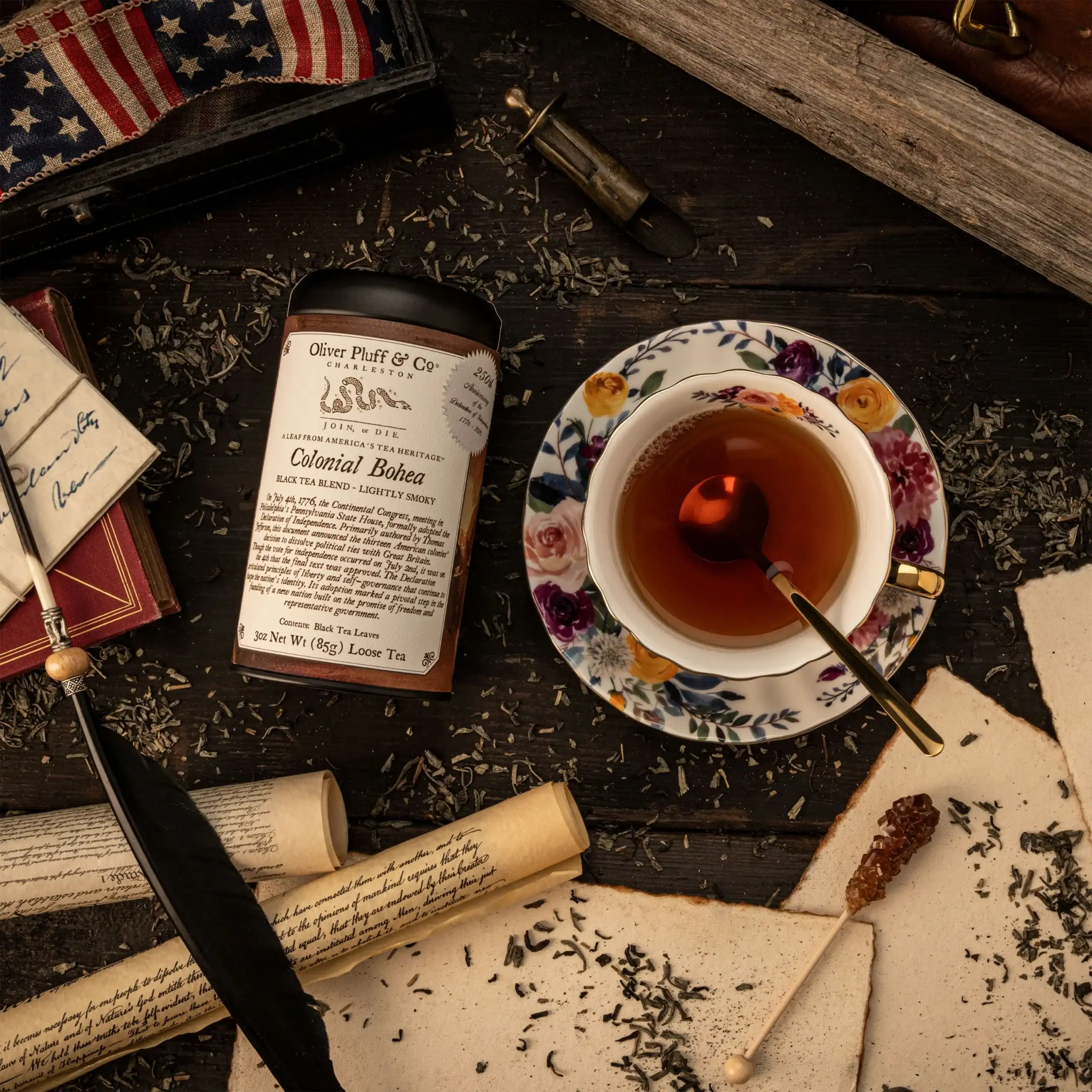 A floral teacup filled with tea sits on a dark table surrounded by loose tea leaves, a tea tin, an old quill, antique papers, cinnamon teacakes, and an American flag pouch-evoking the elegant charm of Dolley Madison.