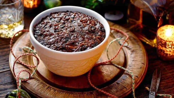 A bowl of Christmas pudding, prepared on Stir-Up Sunday, sits on a wooden board surrounded by festive decorations, candles, a drink, scissors, string, and holiday cards.