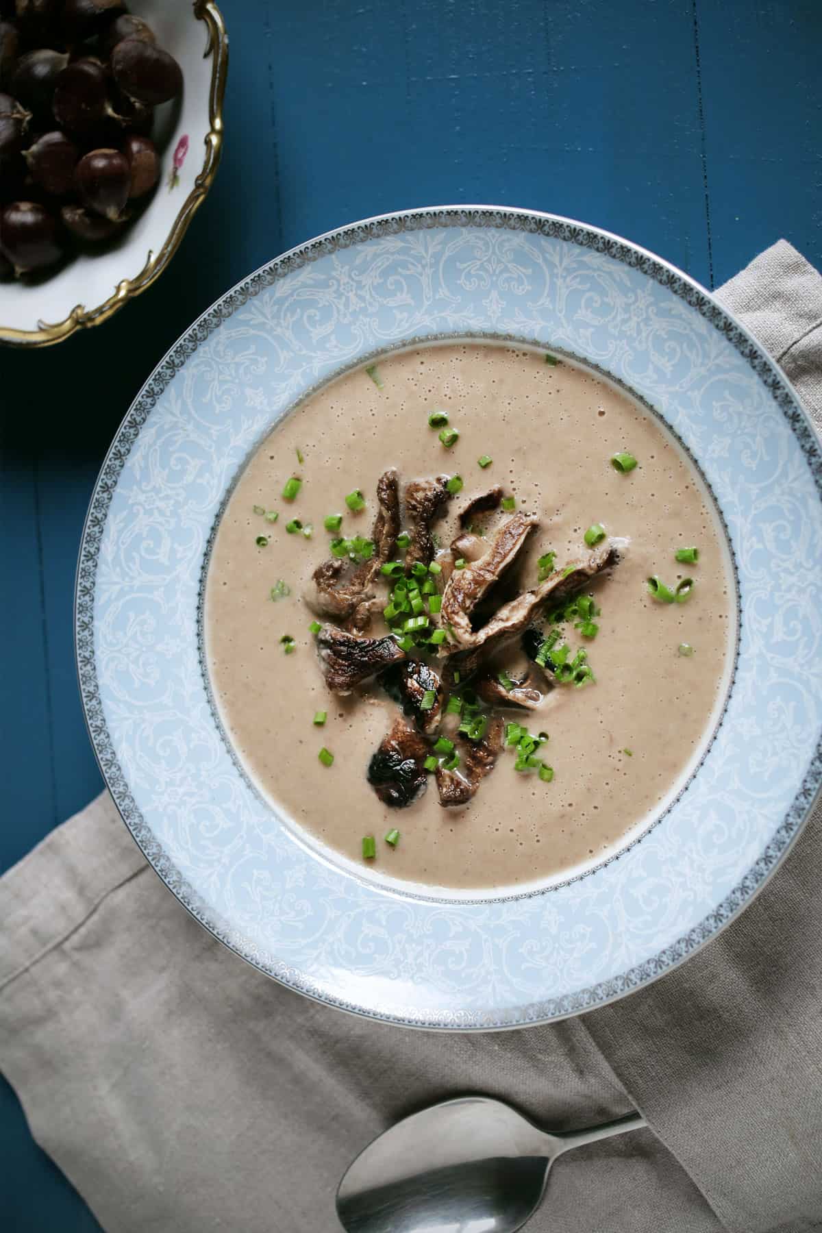 Vertical view of a bowl of creamy chestnut soup with a silver spoon and linen nearby and a bowl of roasted chestnuts.