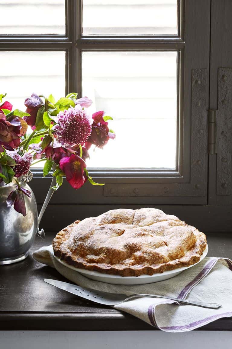 A Thanksgiving Pie sits on a folded cloth beside a knife and a metal vase of flowers on a windowsill, bathed in soft light coming through the window.