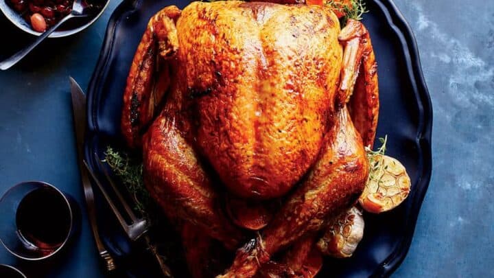 A whole roasted turkey on a black platter with herbs, roasted garlic, two glasses of red wine, and a bowl of olives on a blue surface—perfect inspiration for Turkey recipes for Thanksgiving.