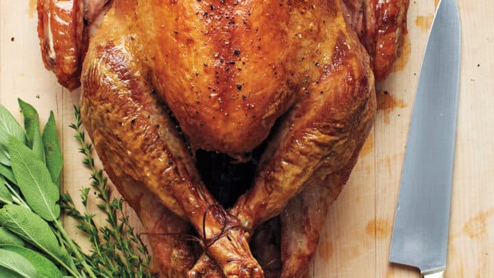 A whole roasted turkey on a wooden cutting board next to a chef's knife, fresh herbs, and grapes—perfect inspiration for Turkey recipes for Thanksgiving.