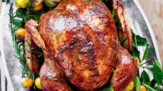 Roast turkey on a large platter, garnished with fresh herbs and assorted small citrus fruits, is the perfect centerpiece for Turkey recipes for Thanksgiving, with carving utensils beside the dish on a wooden table.