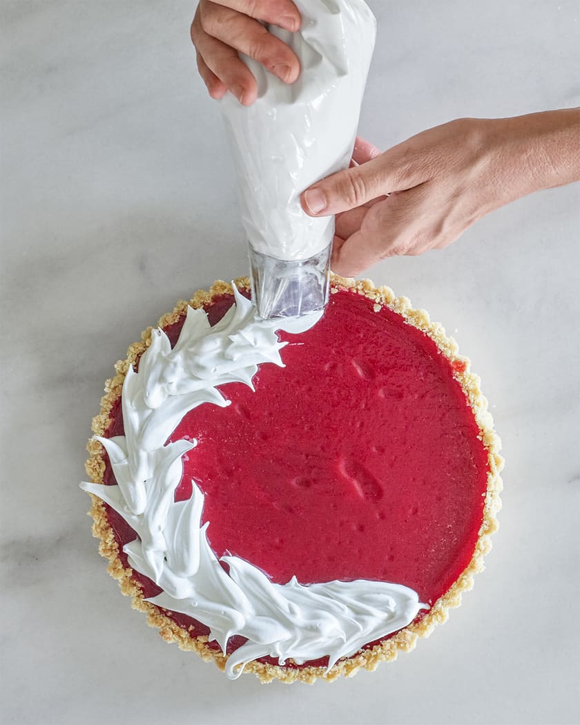 A hand pipes white whipped cream onto the edge of a Thanksgiving Pie-inspired red fruit tart on a marble surface.