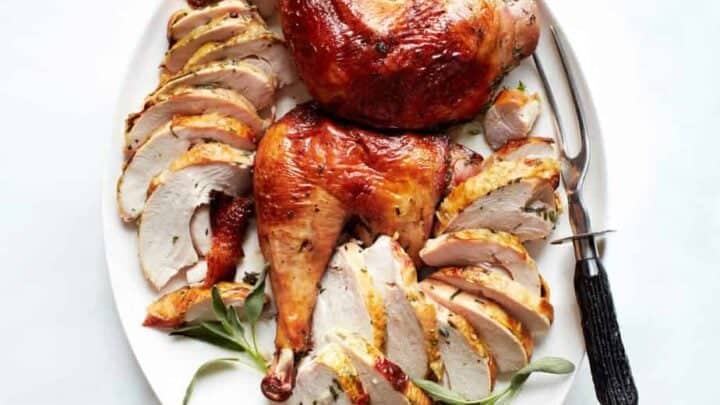 Sliced roasted turkey breast and whole turkey legs arranged on a white platter with fresh herbs, accompanied by a carving fork and knife—perfect for those seeking classic turkey recipes for Thanksgiving.