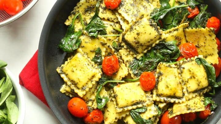 A skillet filled with ravioli pasta, spinach, cherry tomatoes, and pesto—perfect for Healthy 15 Minute Dinners—sits next to a bowl of fresh spinach and a colander of cherry tomatoes.
