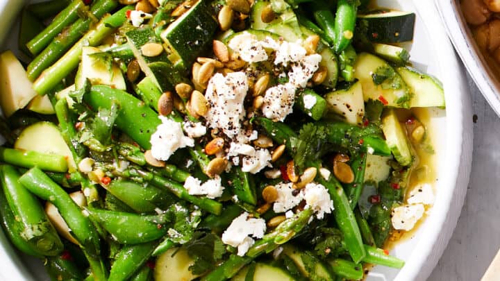 A bowl of green vegetable salad with cucumbers, green beans, snap peas, crumbled cheese, herbs, and seeds, topped with cracked black pepper—perfect for a fresh side at a small Thanksgiving dinner.