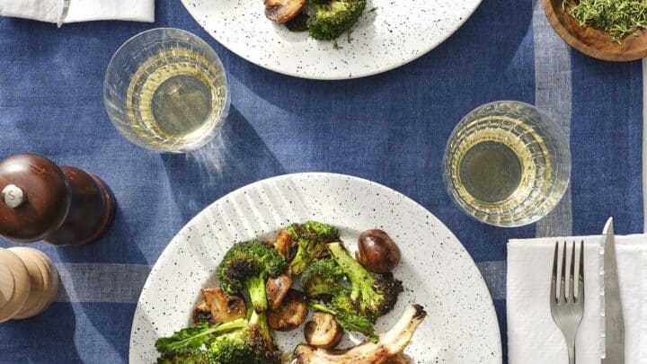 Two plates with pork chops topped with melted cheese, roasted broccoli, and mushrooms make perfect healthy fall dinners, set on a white table with blue placemats and glasses of white wine.