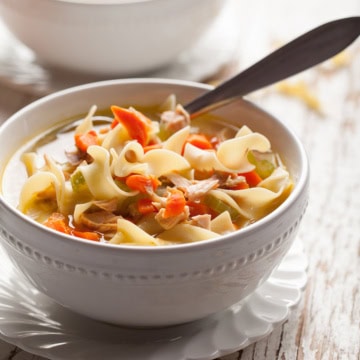 Bowl of turkey noodle soup with a spoon resting on a white scalloped plate.